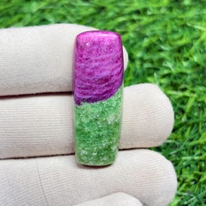 Könnte beinhalten: Ein rechteckiger Edelstein mit polierter Oberfläche. Die obere Hälfte ist ein leuchtendes Magenta mit Wirbelmustern, das in eine strukturierte grüne Basis übergeht. Der Edelstein wird vor einem weißen Stoffhintergrund gehalten.