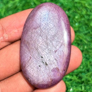 May include: A polished, oval gemstone in pink and purple hues. The stone features a textured surface with fine, intersecting lines. The gemstone is held in a hand against a blurred green background.