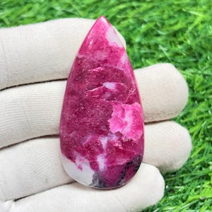 May include: A teardrop-shaped gemstone with a vibrant mix of colors. The stone features a deep magenta hue with white and black inclusions. The polished surface reflects light, highlighting the natural patterns and textures. The gemstone is held against a white background.