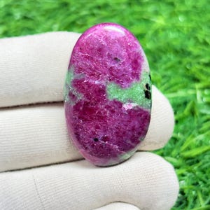 May include: Oval-shaped ruby zoisite gemstone. The polished stone features a vibrant mix of deep pink and green hues, with a smooth, glossy surface. The gemstone is held against a backdrop of green grass.