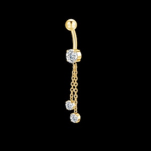 May include: A gold-coloured belly button ring featuring three clear, round gemstones suspended from a fine chain. The top of the ring has a small ball, and the gemstones are set in gold-coloured bezels. The background is black.