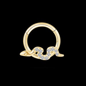 Può includere: Un anello per il naso incernierato color oro con un design a forma di serpente. Il corpo del serpente è adornato da gemme chiare e scintillanti. L'anello è posto su uno sfondo nero.