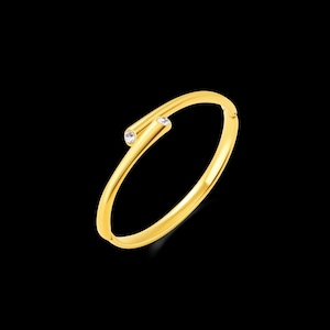 May include: A gold-coloured bangle bracelet with a minimalist design. The bracelet features a slender band that curves around and has a small, clear gemstone set into a cylindrical accent. The bracelet is set against a black background.