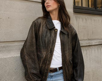 May include: An oversized, distressed dark brown leather bomber jacket with a classic collar, long sleeves, ribbed cuffs, and a front zipper closure. The jacket features a worn-in texture, two side pockets, and a single visible gold-tone metal snap button.