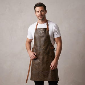 Könnte beinhalten: Braune Lederschürze mit großer Fronttasche und verstellbaren Trägern. Auf der Schürze steht "Custom Logo" auf der Brust. Die Schürze wird über einem weißen T-Shirt getragen.