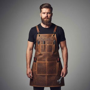 Pode incluir: Avental de couro castanho com vários bolsos e porta-ferramentas. O avental tem a palavra "Customization" em relevo na parte superior. Ferramentas, incluindo chaves e chaves de fenda, são visíveis nos bolsos. O avental tem alças de couro e ferragens de latão.