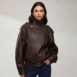 Könnte beinhalten: Braune Kunstlederjacke mit hohem Kragen und Reißverschluss. Die Jacke hat einen Gürtel in der Taille und ein Knopfdetail an der Schulter. Das Model trägt eine blaue Jeans.