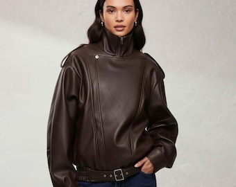 May include: A brown faux leather jacket with a high collar and a zip-up front. The jacket features a belt at the waist and a button detail on the shoulder. The model is wearing blue jeans.
