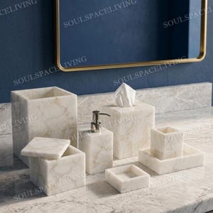 Könnte beinhalten: Ein Badezimmer-Set aus Marmor mit Seifenspender, Taschentuchbox, kleiner Ablage und anderem Zubehör. Das Set hat ein weißes und graues Adernmuster. Die Artikel sind auf einer Marmor-Arbeitsplatte mit einem goldgerahmten Spiegel angeordnet.