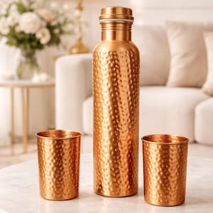 May include: A copper-colored water bottle and two matching tumblers with a hammered texture. The bottle has a screw-on cap. The items are displayed on a marble-topped table with a gold base. The background includes a sofa and flowers.