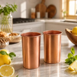 Könnte beinhalten: Zwei zylindrische Kupferbecher mit einer glatten, metallischen Oberfläche. Die Becher stehen aufrecht und haben ein schlichtes Design mit geraden Wänden und einem dünnen Rand an der Oberseite. Die Kupferoberfläche reflektiert das Licht.