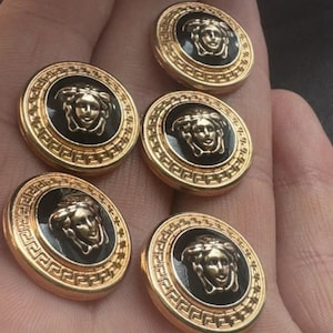 Puede incluir: Seis botones dorados con un centro negro. Cada botón presenta un diseño de cabeza de Medusa y un borde de llave griega. Los botones son redondos y tienen un acabado brillante, adecuados para ropa o proyectos de artesanía.