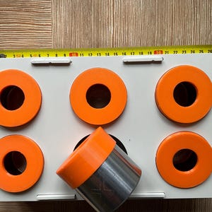 Könnte beinhalten: Eine weiße rechteckige Ablage mit sechs orangefarbenen kreisförmigen Komponenten und einer orangefarbenen und silbernen zylindrischen Komponente. Ein gelbes Maßband liegt oben auf, mit Messungen in Zentimetern.
