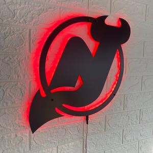 May include: A black metal sign displaying the New Jersey Devils logo, a stylized "N" within a circle, with devil horns. The sign is illuminated with a vibrant red LED glow around the logo's outline, creating a striking visual effect.