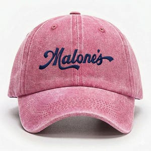 May include: A pink baseball cap with a washed, distressed texture. The front features the word Malone's embroidered in dark navy blue script lettering. The cap has a curved brim with visible stitching and a structured crown with eyelet vents.