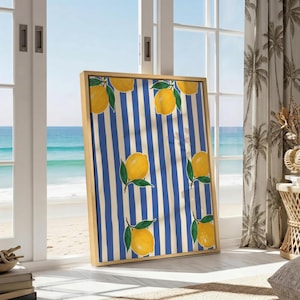 May include: A framed artwork featuring a pattern of yellow lemons with green leaves on a blue and white striped background. The artwork is displayed indoors near a window overlooking the ocean. The frame is a light wood color.