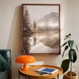 May include: A rectangular framed wall art print depicting a misty mountain landscape with a lake, pine trees, and snow-capped peaks. The scene features muted brown and grey tones, reflecting the mountains and trees in the calm water surface.