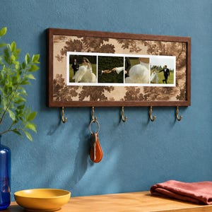 Könnte beinhalten: Ein rechteckiger Schlüsselhalter zur Wandmontage mit einem dunklen Holzrahmen und einer zentralen Platte mit braun-beigem Toile-de-Jouy-Muster. Die Platte zeigt vier horizontale Hochzeitsfotos und verfügt über fünf messingfarbene Metallhaken.