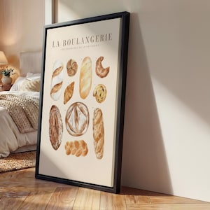 Pode incluir: Uma impressão emoldurada com ilustrações em aquarela de vários produtos de panificação, incluindo baguetes, croissants e pãezinhos. O texto "LA BOULANGERIE" e "LES ESSENTIELS DE LA PATISSERIE" é exibido na parte superior. A obra de arte tem uma paleta de cores neutras.