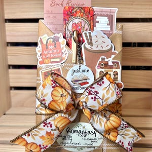 Puede incluir: Un set de regalo de libro envuelto con una cinta con estampado de calabazas, un llavero circular con el texto just one more chapter y varias pegatinas que incluyen una taza de cacao, una librería y una tarjeta de reseña de libro con la etiqueta Romantasy.
