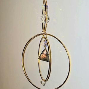 May include: A gold-colored suncatcher with a geometric design. It features a chain, three gold rings, and several crystal accents. A triangular stone and a large, faceted crystal are suspended within the rings, creating a decorative and reflective piece.