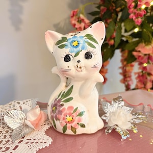 Puede incluir: Una figura de gato de cerámica blanca con detalles florales pintados a mano en azul, rosa y verde. El gato tiene grandes ojos negros, pestañas largas y una boca pequeña. Está decorado con una flor azul en la frente y flores rosas en el costado.