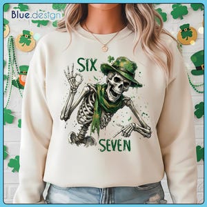 Op de afbeelding: Crèmekleurige sweatshirt met een skelet met een groene hoed en sjaal, met de groene woorden "SIX" en "SEVEN". Het skelet maakt een "okay" handgebaar. Het ontwerp heeft een St. Patrick's Day thema.