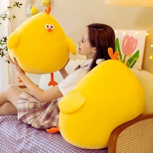 May include: Two large, plush yellow chick-shaped pillows. One is held up, showing orange feet, a beak, and eyes. The other is resting on a bed. The pillows have small wings and orange accents. The background includes a bed with a blue and white checkered pattern.