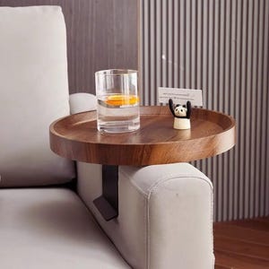 May include: A round, dark brown wooden sofa arm tray with a clamp. A clear glass with a slice of orange sits on the tray, along with a small panda figurine holding a card. The tray is attached to a light gray sofa arm.