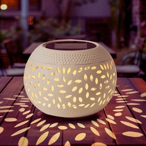 Può includere: Una lanterna solare sferica color crema con una parte superiore strutturata e ritagli a forma di foglia. La lanterna è appoggiata su un tavolo di legno, proiettando motivi di luce. La parte superiore ha un pannello solare.