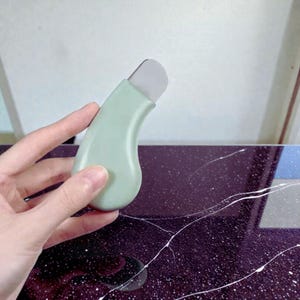 May include: A light green facial skin spatula with a silver blade. The spatula has a curved handle and is held by a hand. The background is a light purple surface with white streaks.