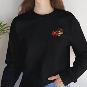 Puede incluir: Sudadera negra con un pequeño diseño bordado de un guante de boxeo y la cara de un mono. El diseño es en tonos rojos, marrones y beige. La sudadera es de cuello redondo.