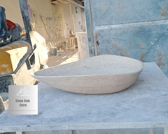 Puede incluir: Un gran lavabo de piedra beige con forma ovalada y lisa. El lavabo se exhibe sobre una superficie gris, con un logotipo de Stone Sink Store visible en primer plano. El lavabo parece estar hecho de piedra natural.