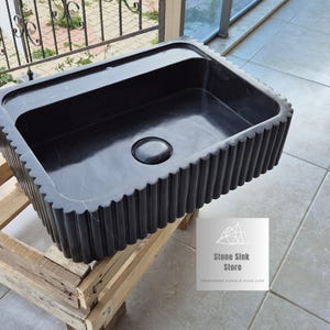 May include: A black rectangular stone sink with a fluted edge and a central drain. The sink is displayed on a wooden pallet. The Stone Sink Store logo is visible.