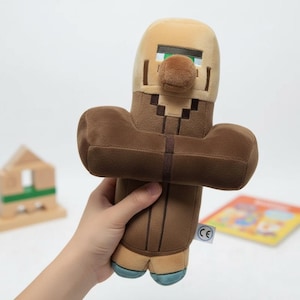 Pode incluir: Um brinquedo de pelúcia Minecraft Villager em tons de castanho, bege e azul. O brinquedo tem um design em blocos com um nariz grande e olhos verdes. O brinquedo é segurado na mão de uma pessoa.
