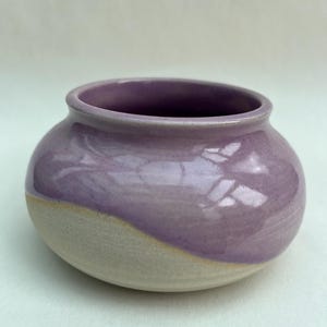 May include: A small, round ceramic bowl with a wide opening and a smooth, glossy lavender glaze on the upper portion. The lower section of the bowl features an unglazed, matte, off-white clay finish with a curved, diagonal transition line.