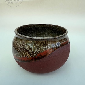 May include: A handmade ceramic bowl with a rustic design. The bowl has a textured, reddish-brown base that blends into a brown and tan upper section with a dark rim. The words "RUSTIC HANDS POTTERY" are printed on the top left.