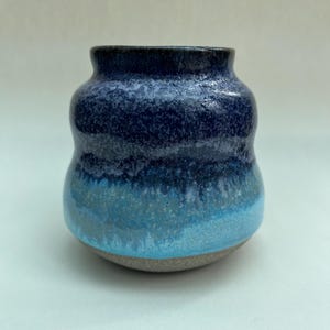 May include: A small ceramic vase with a bulbous shape, featuring a glossy glaze that transitions from a deep dark blue at the top to a vibrant turquoise in the middle, ending in a raw, unglazed light grey base with a visible speckled texture.