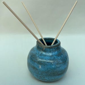 May include: A small, round, ceramic diffuser vessel with a mottled blue and dark grey glaze. Three thin, light-coloured wooden reeds are inserted into the narrow opening at the top of the pot, extending outwards in different directions.