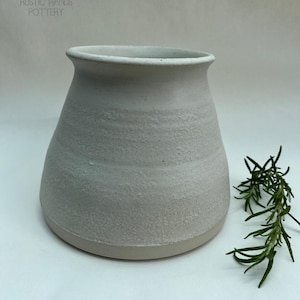 May include: A handmade, off-white ceramic vase with a textured finish. The vase has a broad base, narrowing upwards to a slightly flared rim. A sprig of green rosemary is placed to the right of the vase. The text "RUSTIC HANDS POTTERY" is visible.