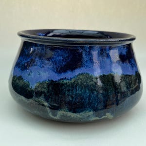 May include: A ceramic bowl with a wide, rounded body and a flared rim. The exterior features a glossy glaze with a deep blue upper section that transitions into a mottled blend of dark blue, forest green, and black tones across the lower surface.