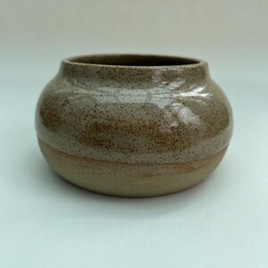 May include: A small, round ceramic bowl with a wide opening. The upper portion features a glossy, speckled beige glaze, while the lower portion remains in a matte, unglazed tan clay finish. The vessel has a smooth, curved silhouette.