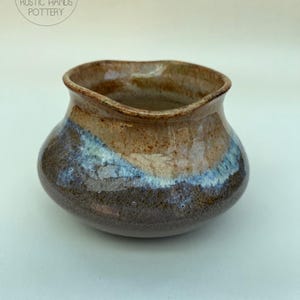 May include: A handmade ceramic bowl with a broad, uneven rim. The bowl displays a mix of colours, including brown, blue, and tan, with a shiny finish. The base is a darker brown, fading to lighter shades towards the top. The logo "RUSTIC HANDS POTTERY" is in the upper left.