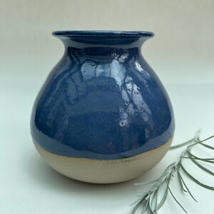 May include: A small, rounded ceramic vase with a glossy, deep blue glazed upper section and a matte, unglazed beige base. The vase features a wide, flared rim and a smooth, bulbous body, showcasing a distinct contrast between the two finishes.
