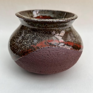 May include: A small, round ceramic pot with a wide, flared rim. The upper portion features a glossy, dark brown and black glaze with speckled textures and a band of deep red. The lower section has a matte, textured, dark maroon finish.