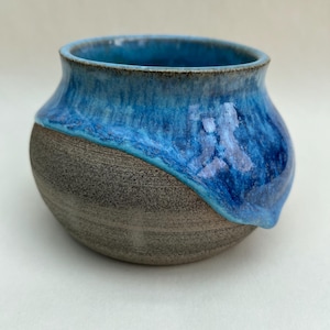 May include: A small, round ceramic bowl featuring a textured, matte grey base and a glossy, vibrant blue glaze that drips down from the rim. The interior of the bowl is finished with the same smooth, deep blue glaze, creating a contrast with the exterior.