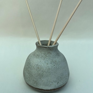May include: A small, round, speckled light grey ceramic vase with a narrow neck and a dark brown base. Three thin, light-coloured wooden reeds are inserted into the opening of the vase, extending upwards in a fan-like arrangement.