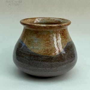 May include: A handmade ceramic vase with a rounded form and a gently widening rim. The vase displays a colour gradient, shifting from a dark brown base to a tan and brown upper section. The text "RUSTIC HANDS POTTERY" is visible.