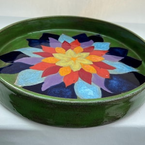 May include: A round ceramic tray with a glossy green exterior and a flat interior surface featuring a vibrant, multi-coloured floral pattern. The design consists of layered, petal-like shapes in shades of yellow, orange, red, purple, and blue.