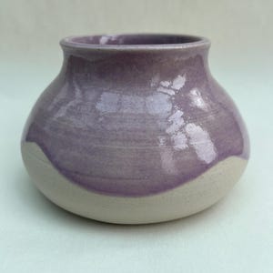 May include: A small, rounded ceramic vase with a wide, curved body and a narrow neck. The upper portion of the vase is coated in a glossy, smooth lavender-purple glaze, while the lower base remains an unglazed, matte, off-white clay finish.